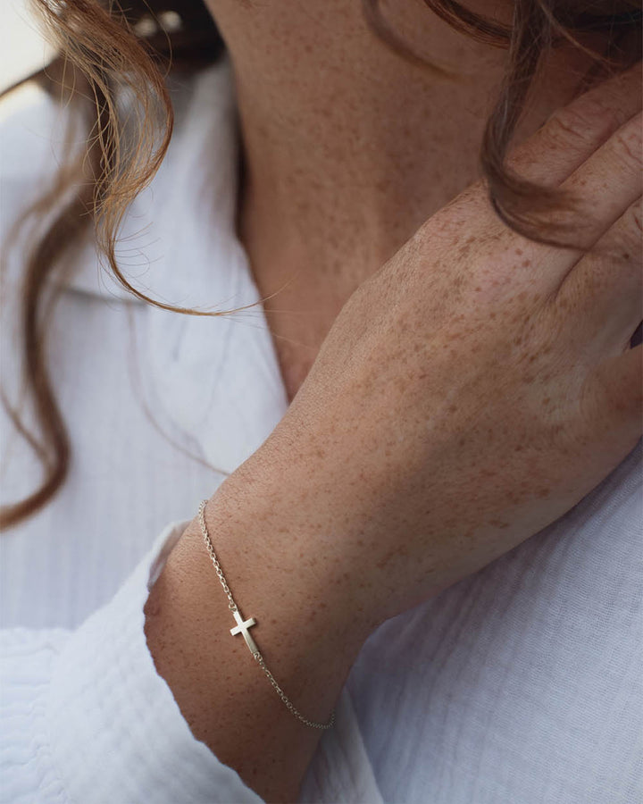 Gold side cross bracelet worn by a woman, hand at the chest